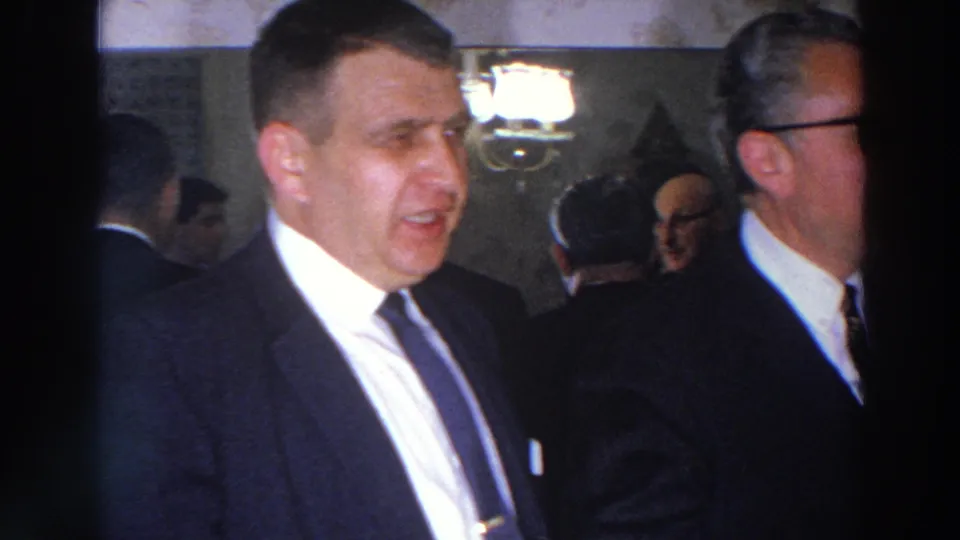 Madison 1966 home-movie frame showing suited men and other adult guests in a dim interior with a chandelier visible behind them.