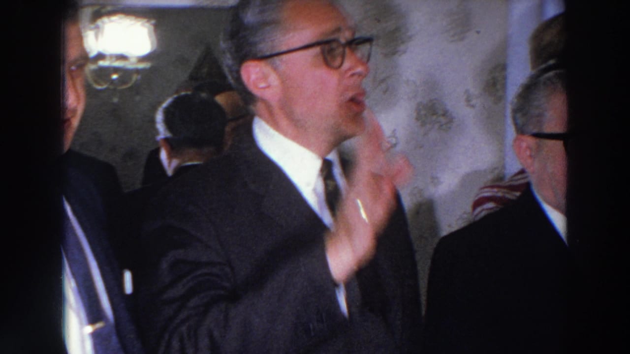 In this 1966 Madison, Wisconsin home-movie frame, a suited man gestures while older adults stand close together inside a crowded room.