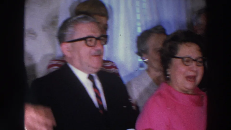A 1966 Madison, Wisconsin frame shows an older suited man and a woman in a bright pink top among adults in an indoor social setting.