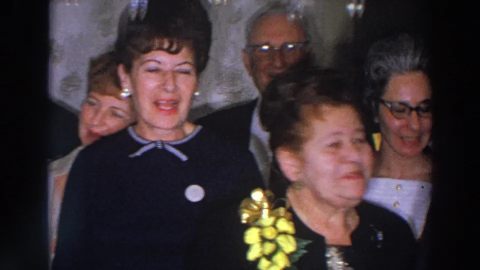 A woman in a dark dress appears mid-song or mid-speech at a 1966 Madison, Wisconsin gathering, with older adults and a yellow corsage nearby.