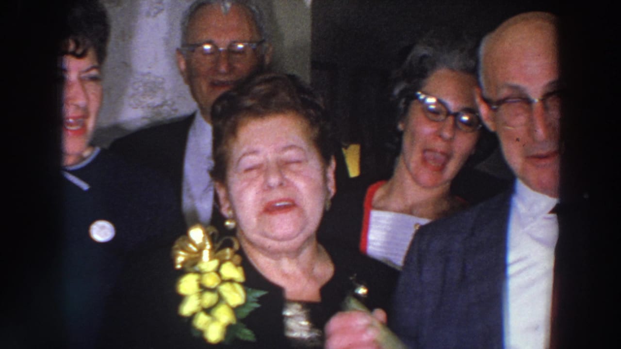 Older adults in formal attire laugh and gather closely in a dim indoor setting.