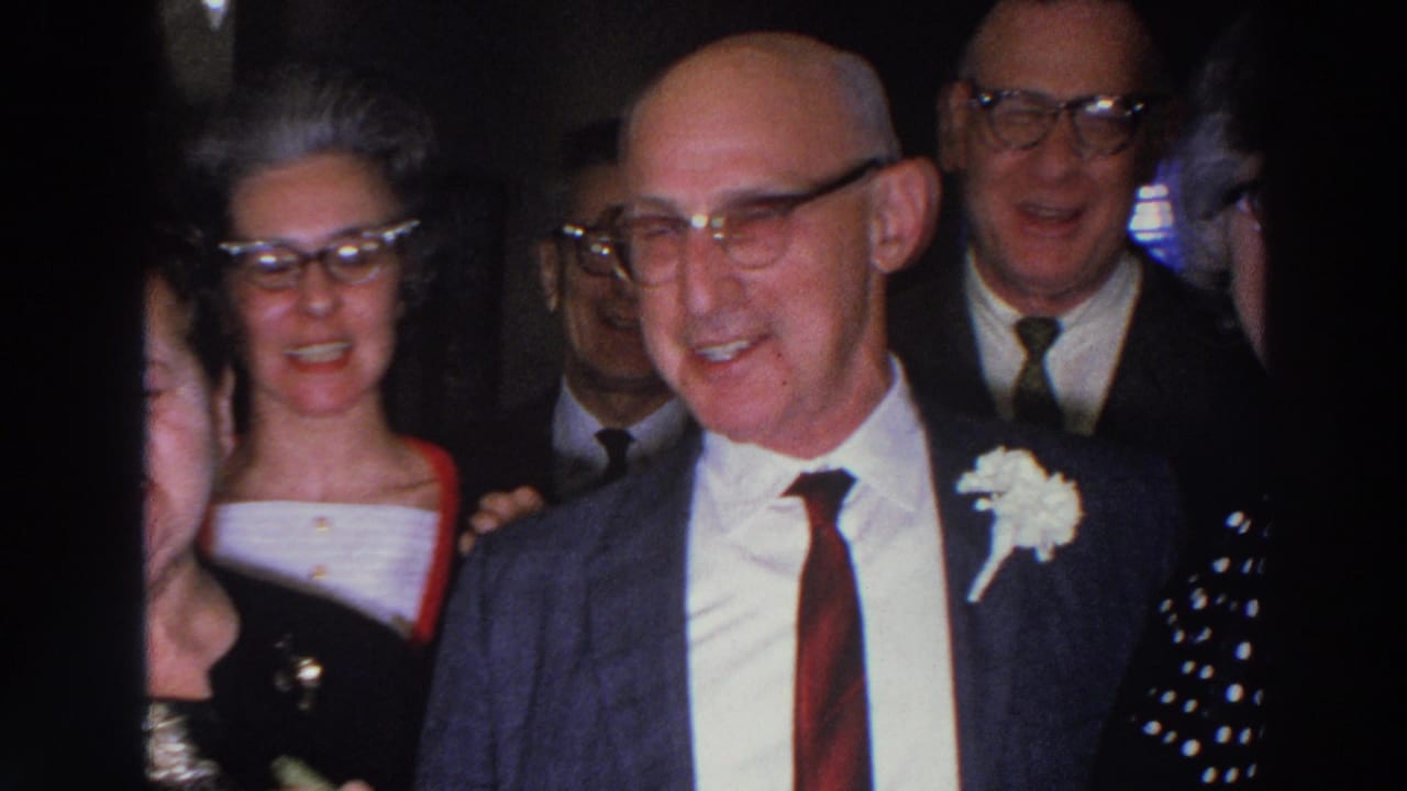 A smiling man in a suit with a boutonniere stands among adults in formal dress.