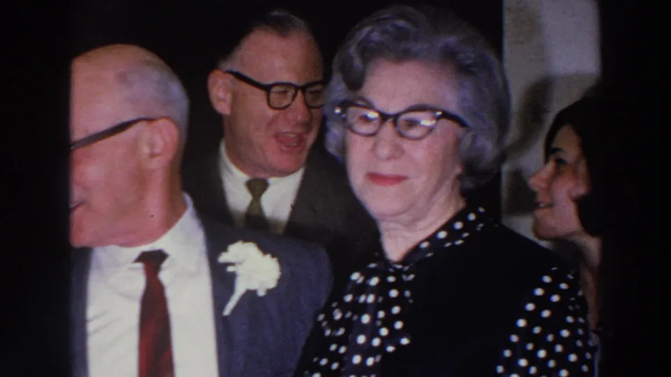 1966 Madison home-movie frame with adults in suits and dresses, including a woman wearing a dark polka-dot dress and glasses.