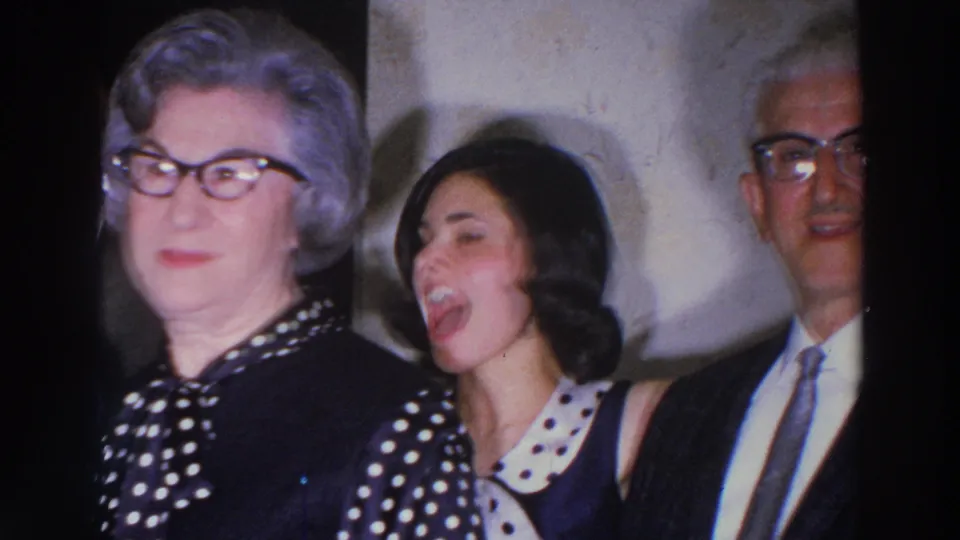 1966 Madison frame of a woman in a polka-dot dress beside a younger woman and a suited man during an indoor social gathering.