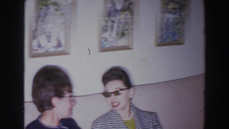 Two women appear seated in a Madison, Wisconsin living room in 1966, one wearing sunglasses and a striped jacket.
