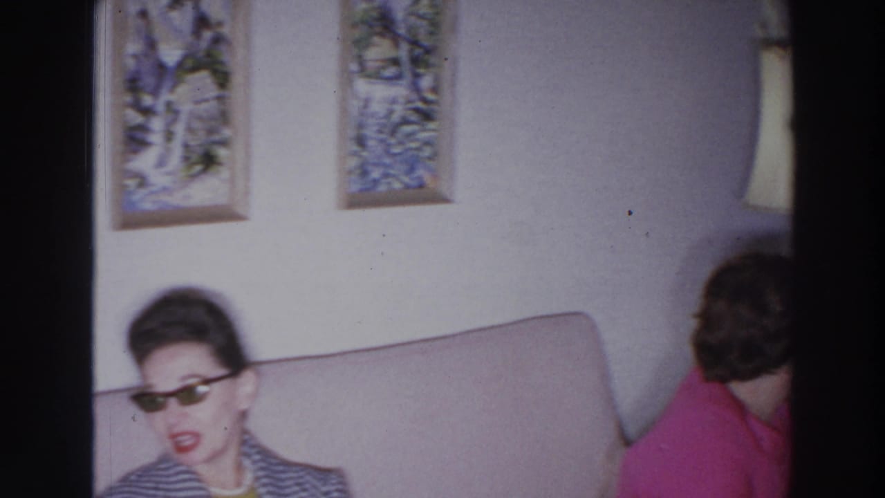 A woman in sunglasses sits on a couch while another woman in pink turns away.