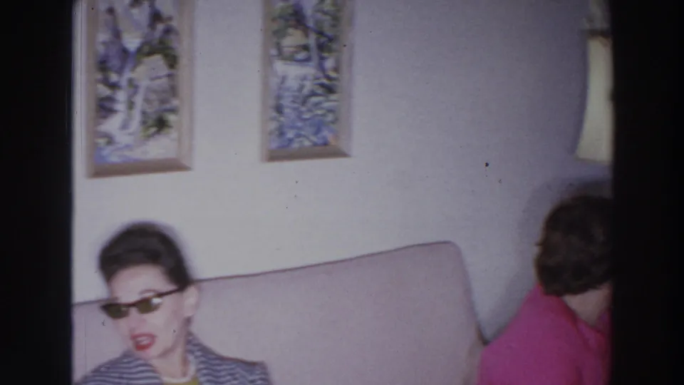 A woman in sunglasses sits on a beige couch as another woman in a bright pink top turns toward the side of the room.
