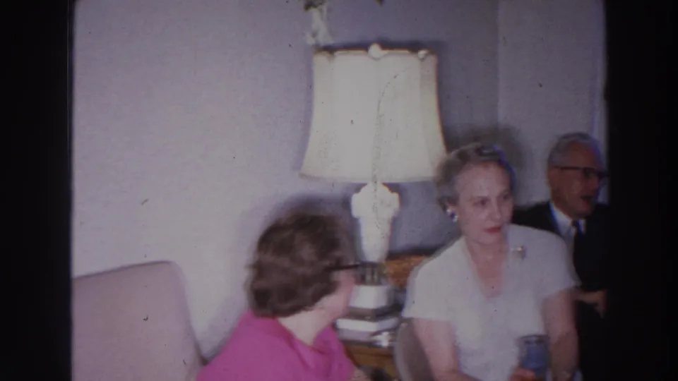 Adults gather in a 1966 Madison living room near a white table lamp, with a man in a dark suit seated behind two women.