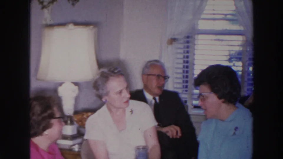 Three women and a man in a dark suit sit near a lamp and curtained window in a Madison, Wisconsin living room.