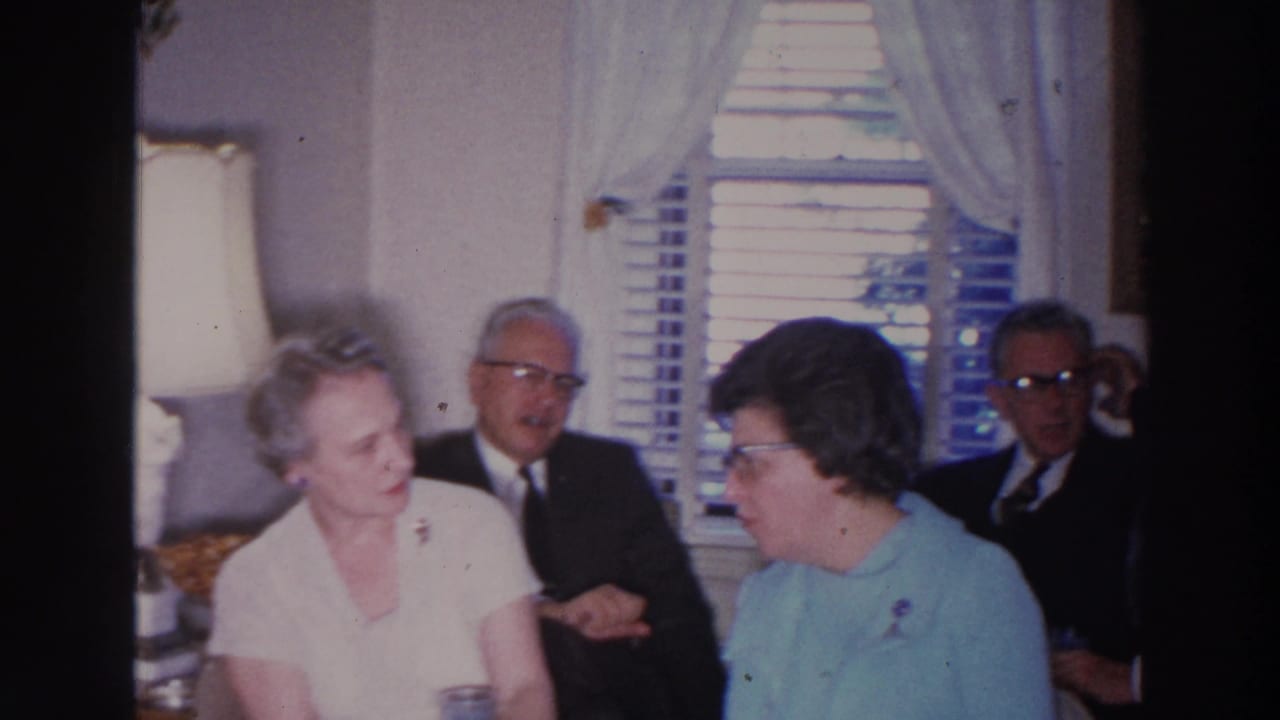 Older adults seated in a living room with a lamp, curtains, and window blinds behind them.