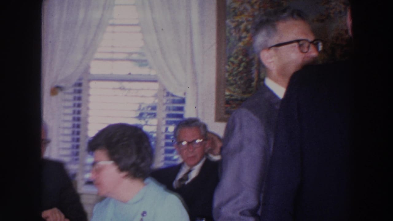 Adults in suits and dresses gather near bright curtained windows inside a living room.