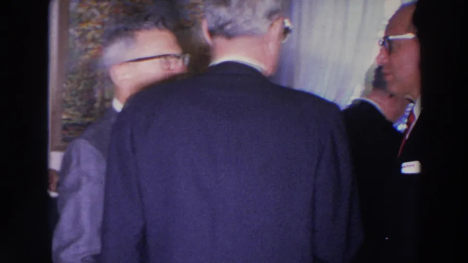 1966 Madison home-movie frame showing suited older men standing and talking in a private living room.