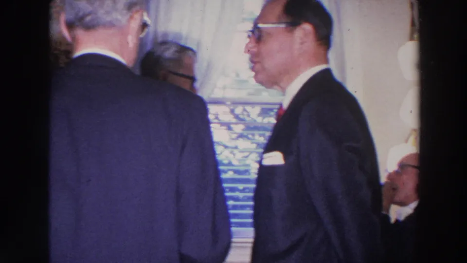 1966 Madison living-room frame with suited men conversing near a curtained window and venetian blinds.
