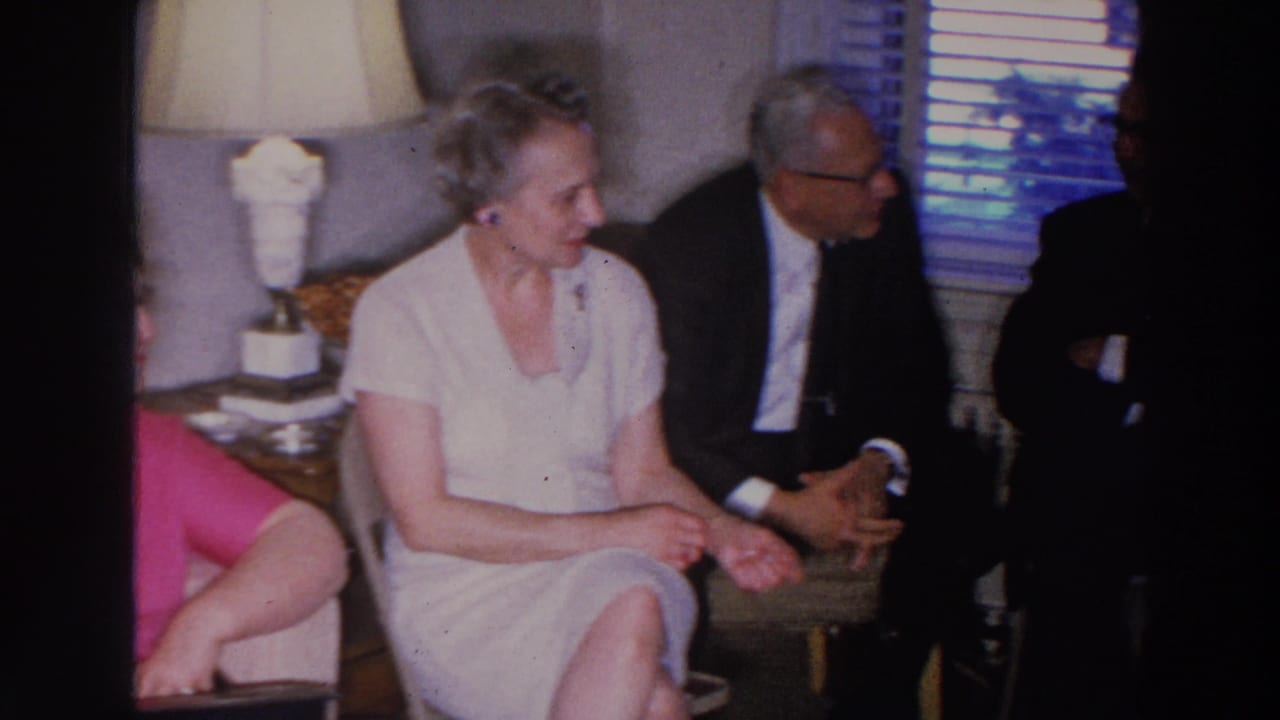 Adults seated in a living room, including a woman in a pale dress and men in suits.