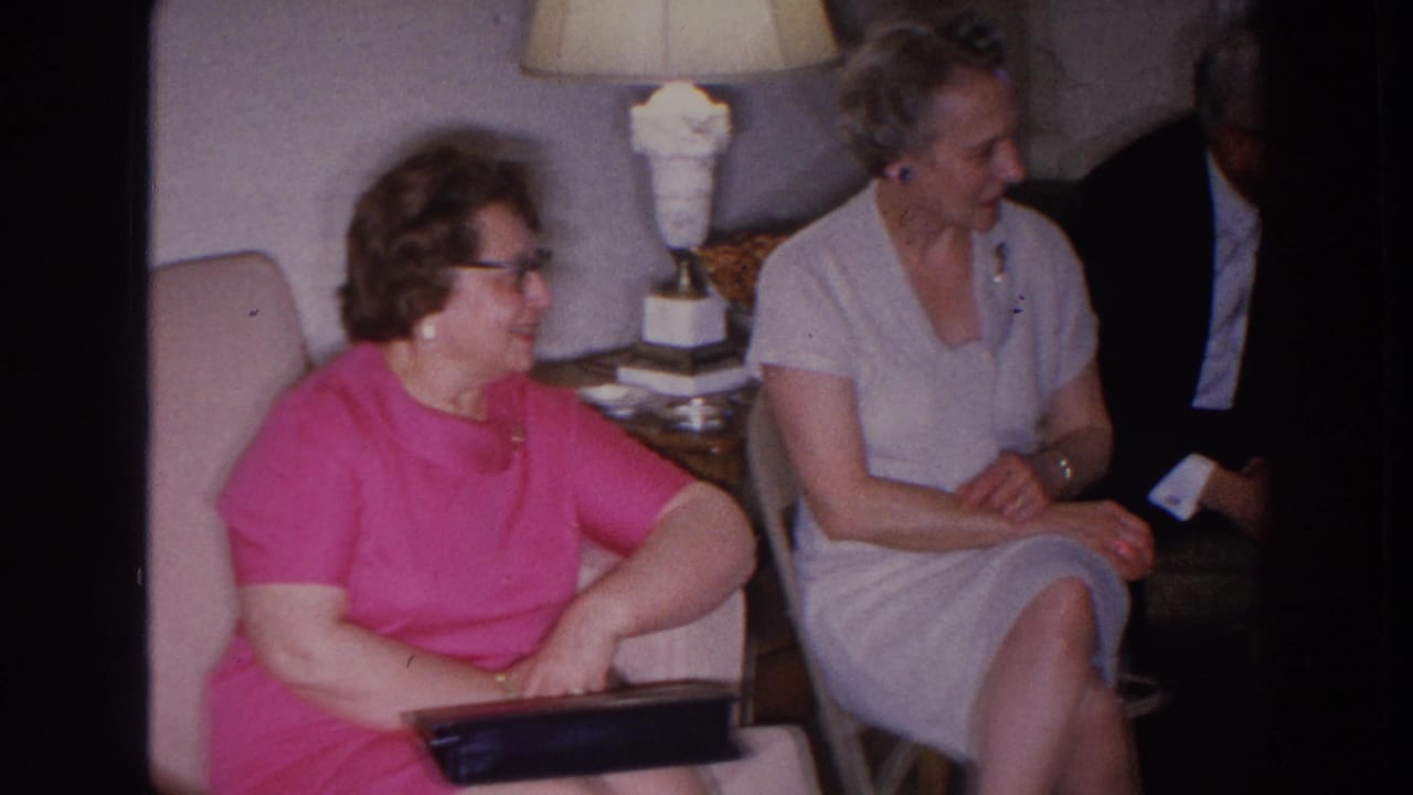 Woman in a pink dress with glasses sits near another woman in a pale dress.