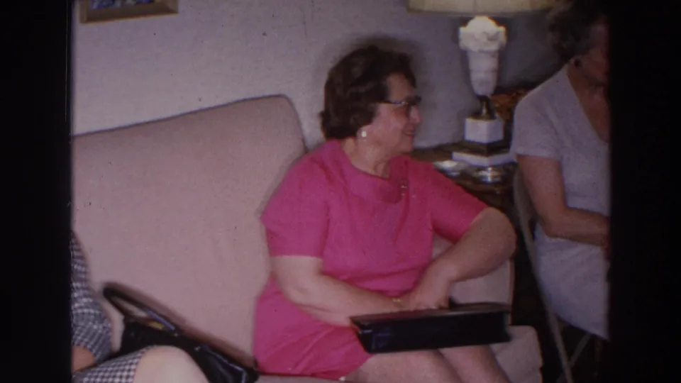 1966 Madison home-movie frame showing a woman in a pink dress seated on a sofa during a living room visit.