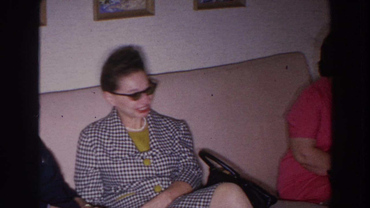 In a 1966 Madison home-movie frame, a woman in sunglasses and a checkered suit sits on a couch during an indoor gathering.