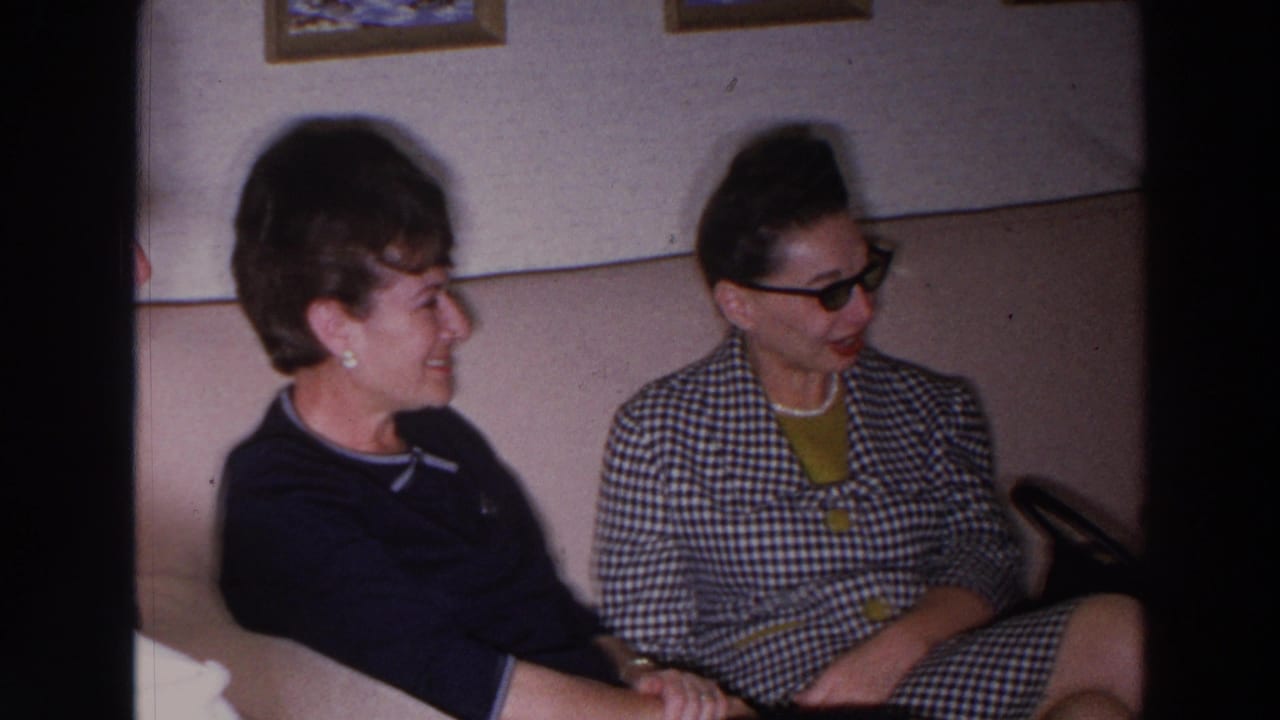 Two women sit close together on a beige couch, smiling and talking in a room.