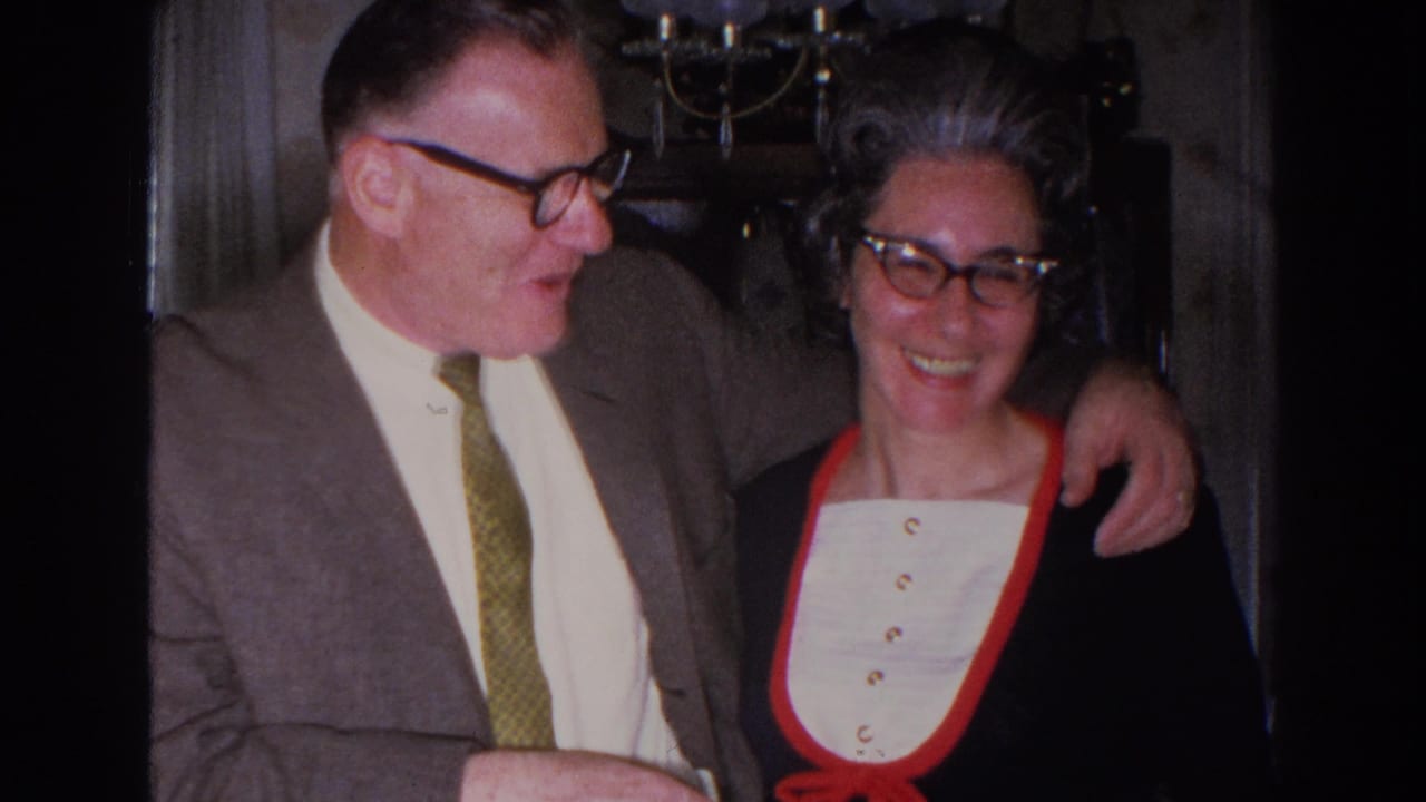 1966 Madison home-movie frame showing a suited man with his arm around a smiling woman in glasses inside a private room.