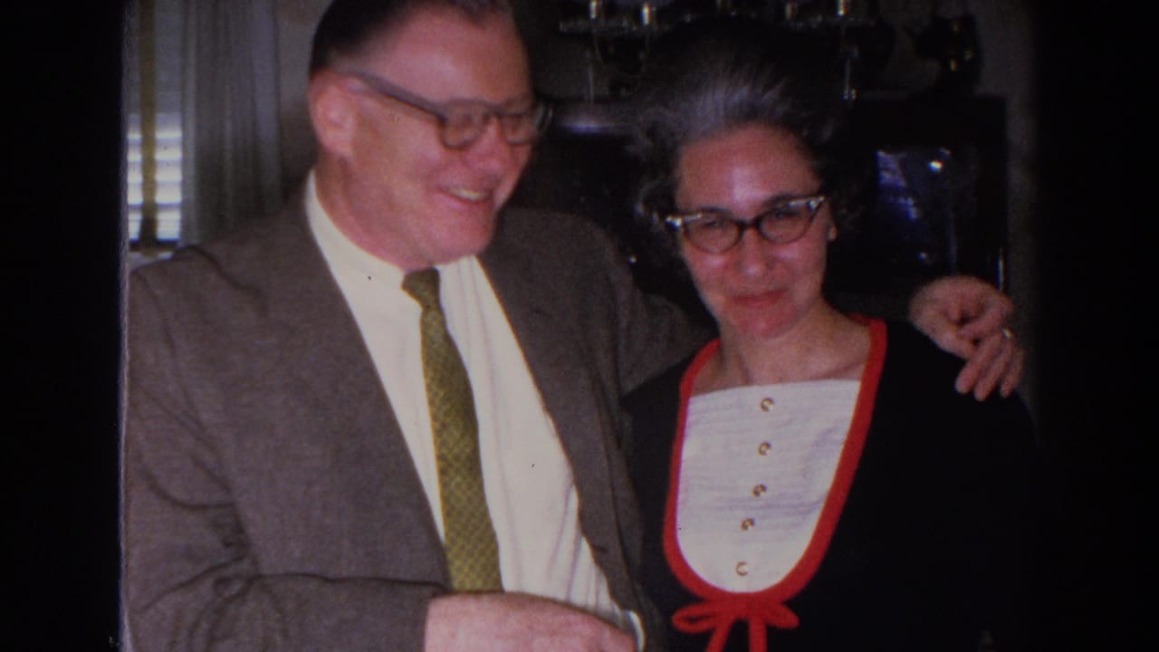 Smiling man in suit stands beside woman in glasses indoors.
