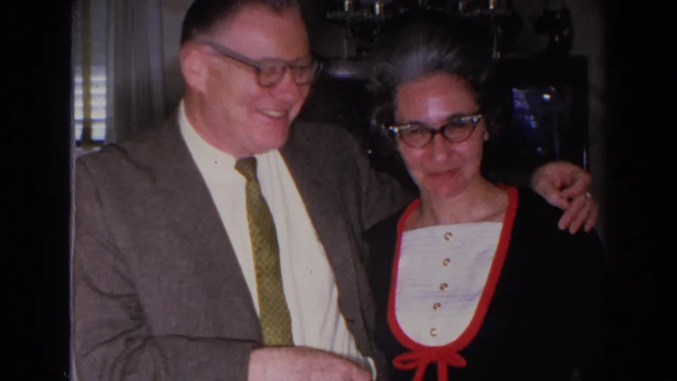 1966 Madison frame of a smiling man in a brown suit standing close to a woman in glasses and red-trimmed clothing indoors.