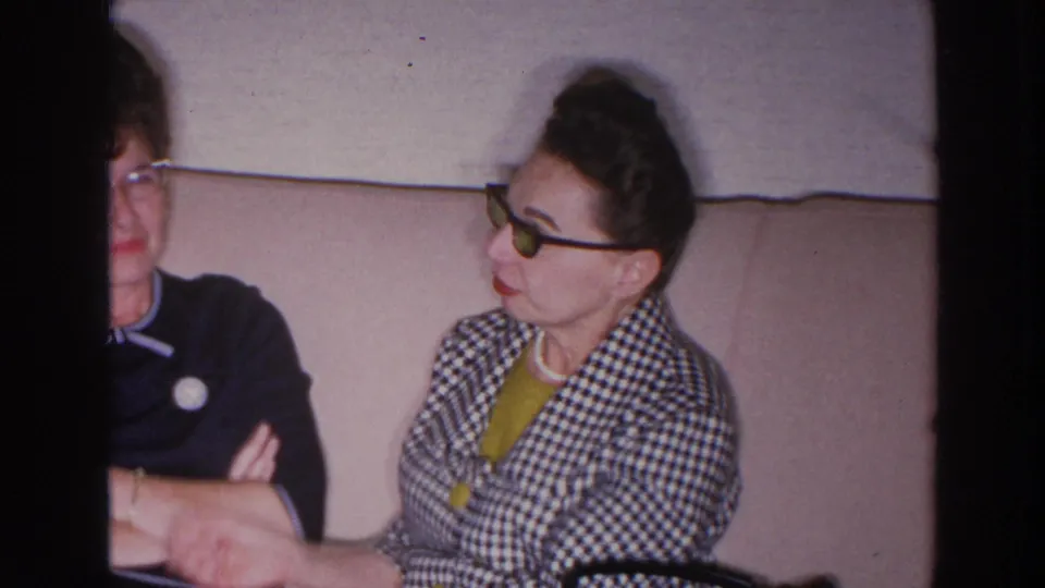 1966 Madison home-movie frame showing two women in glasses seated on a couch, one partly cropped and one in a checkered jacket.