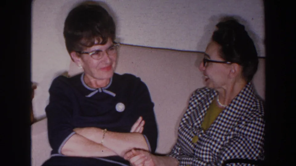 1966 Madison frame of two women seated on a couch, with one wearing a dark bow-collar sweater and the other a checkered jacket.