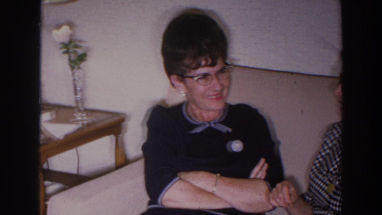 Woman in glasses sits on a tan sofa beside another partially visible person in a living room.
