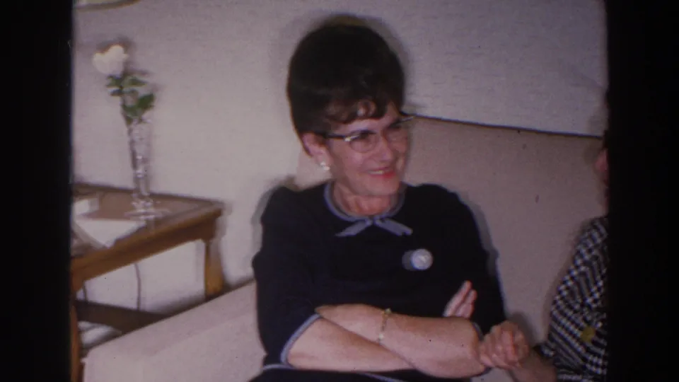 In a 1966 Madison home-movie frame, a woman in glasses and a dark dress sits on a tan sofa near a side table with flowers.