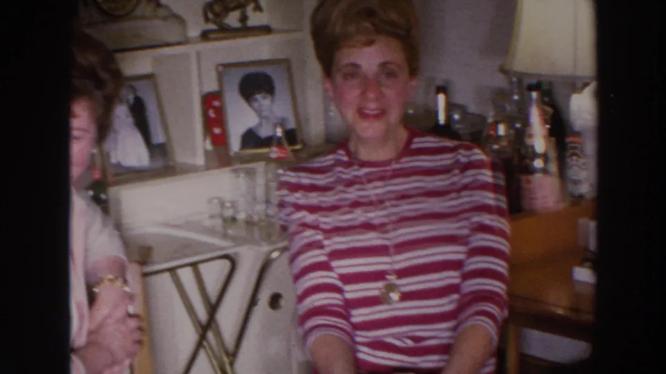 A 1960s Madison home-movie frame shows a woman in a striped sweater seated before framed portraits, shelves, and living-room decor.