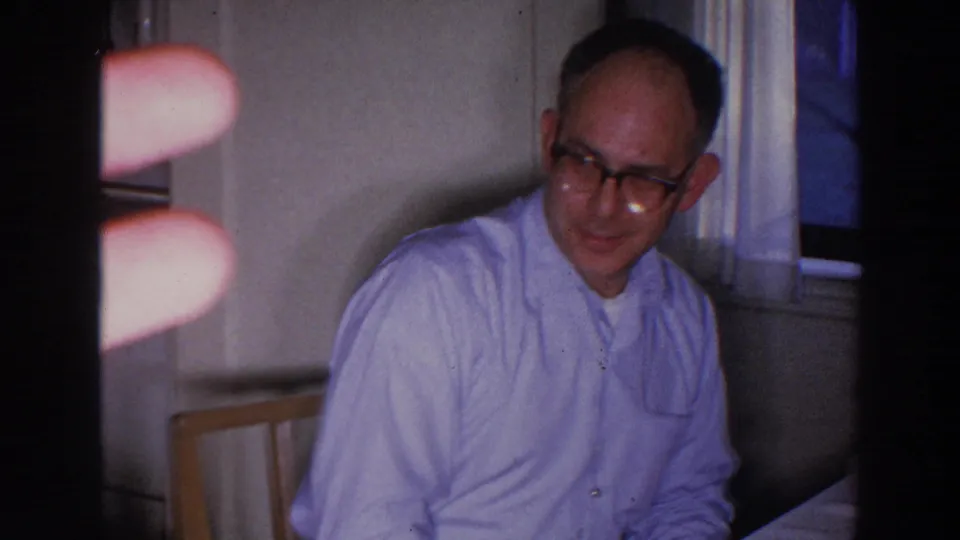 A man in glasses sits in the kitchen, with a finger intruding into the 8mm frame and another person partly visible behind him.