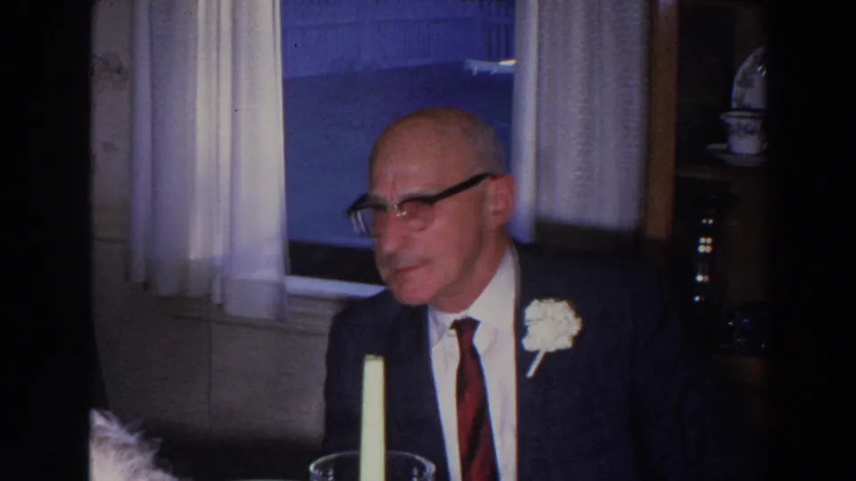 A bald man wearing glasses, a dark suit, and a boutonniere sits at a kitchen table in the 1966 Madison home-movie footage.