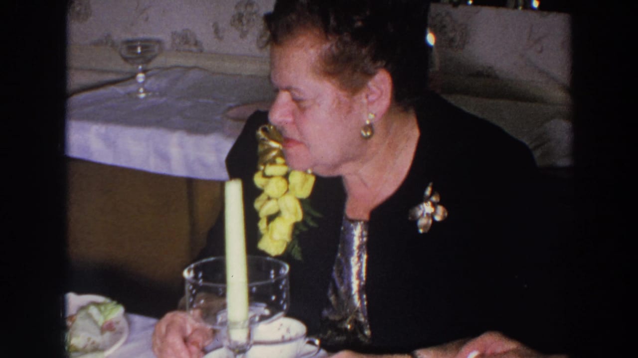 Elderly woman in a black jacket seated at a dinner table with a candle and yellow flowers.
