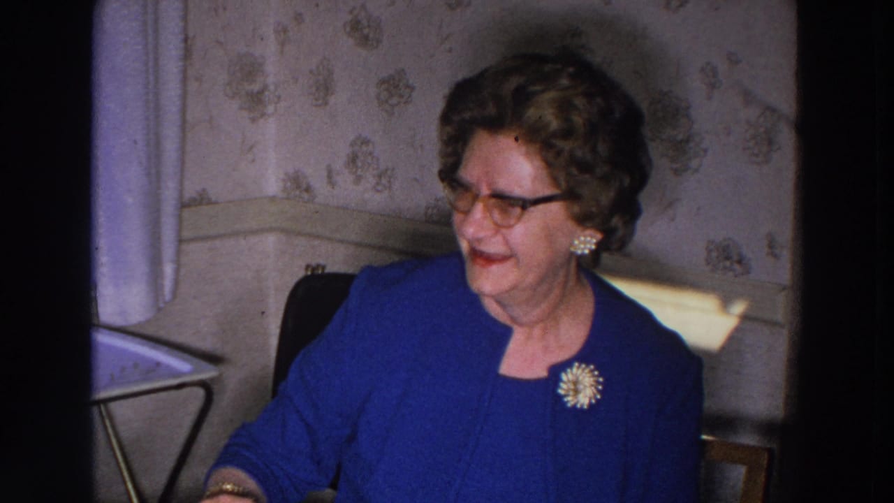 Elderly woman in a blue suit and glasses seated against floral wallpaper.