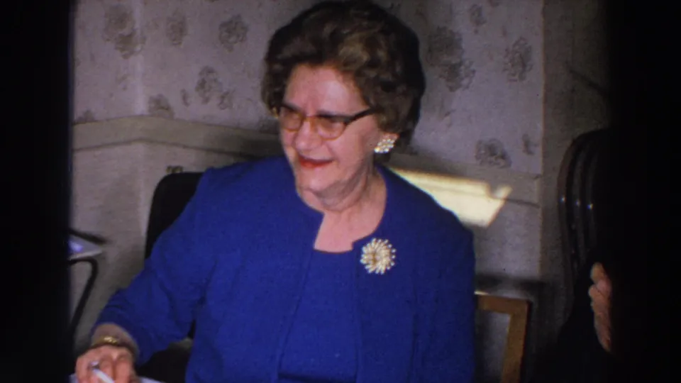 The Madison home-movie footage shows an elderly woman in a blue suit at the table, with floral wallpaper and dining furniture behind her.