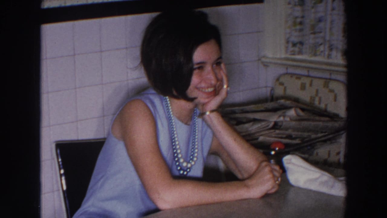 Smiling woman leans on a kitchen counter beside a tiled wall and curtain.