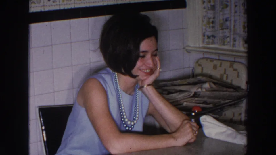 A young woman in a pale blue dress and long pearls looks downward with a smile inside the 1966 home kitchen setting.