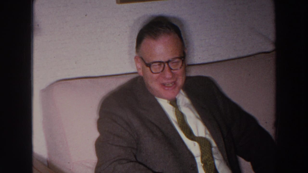 Older man in glasses seated on a light couch, wearing a suit and tie.