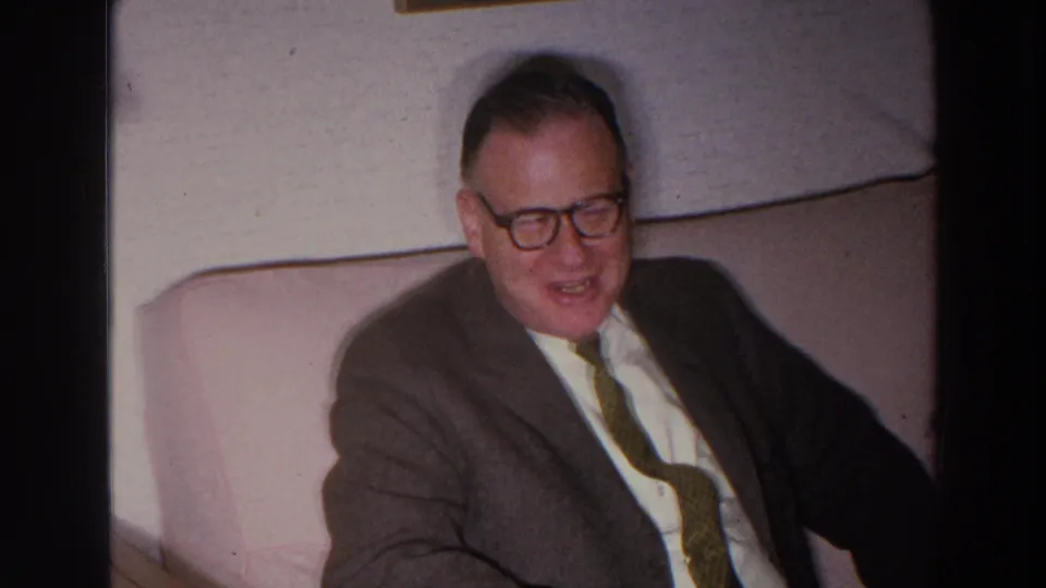 In 1966 Madison, Wisconsin home-movie footage, an older man in glasses sits on a pale couch wearing a brown suit, white shirt, and tie.
