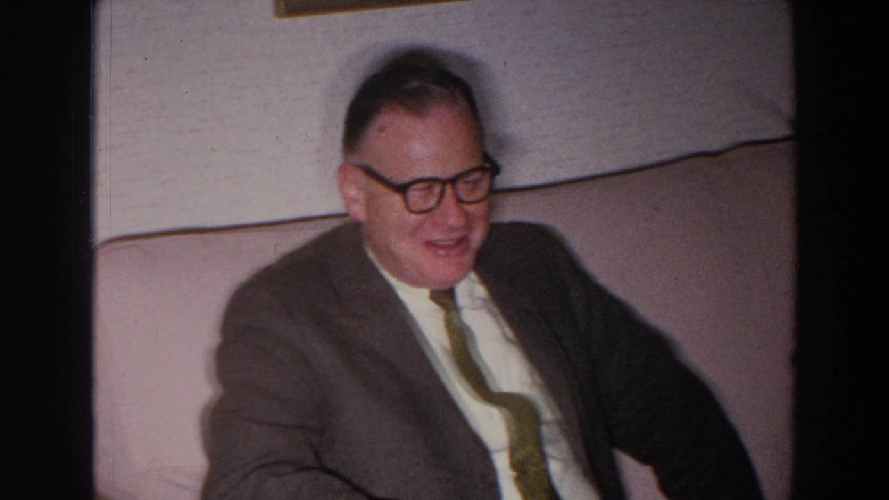 Older man in a suit smiles while seated on a couch indoors.