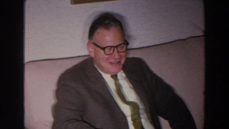 The older man remains seated on the pale couch, smiling in softly lit 1966 Madison, Wisconsin home-movie footage.