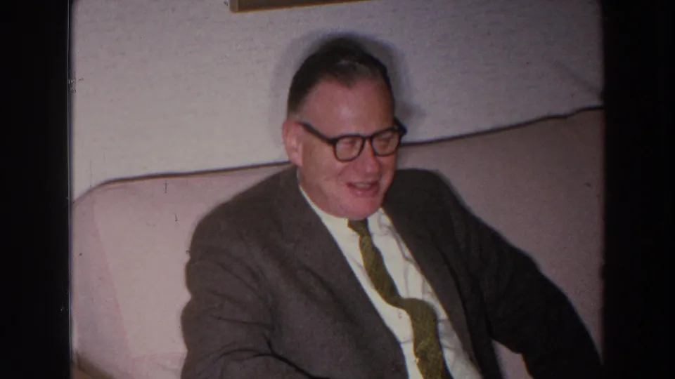 An older man in glasses and a tie looks downward from a light-colored couch in 1966 Madison, Wisconsin home-movie footage.