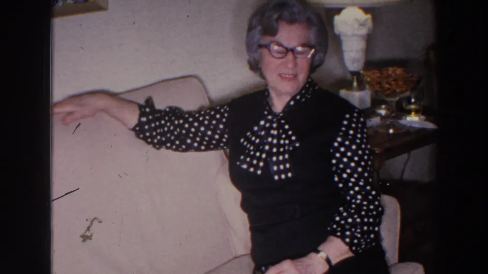 An older woman in glasses and a black polka-dot blouse sits on a pale couch beside a side table in 1966 Madison, Wisconsin footage.