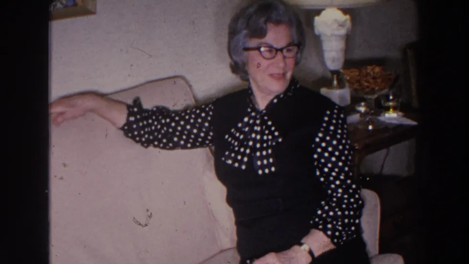 In a 1966 Madison living room, a woman in glasses and a polka-dot blouse sits on a sofa beside a side table and lamp.