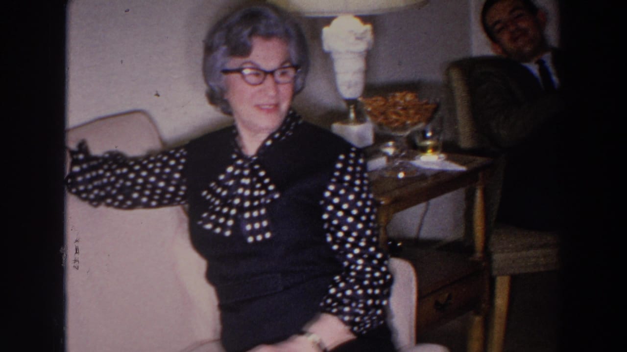 Woman sits on a sofa while a man is partly visible in a chair behind her.