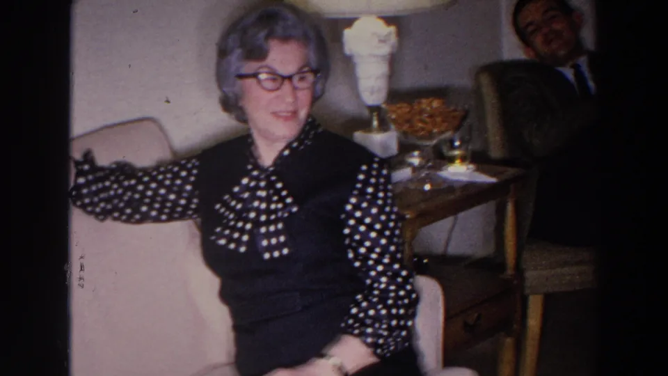 A woman sits on a sofa in a Madison home interior while a suited man is partly visible in the background.
