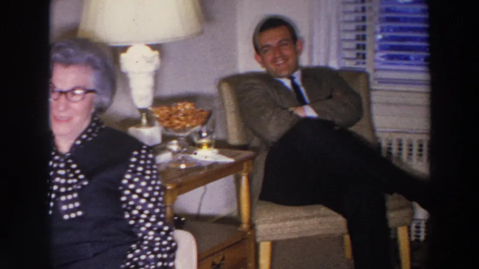 A suited man smiles from a chair in a 1966 Madison living room as the woman remains partly visible at frame edge.