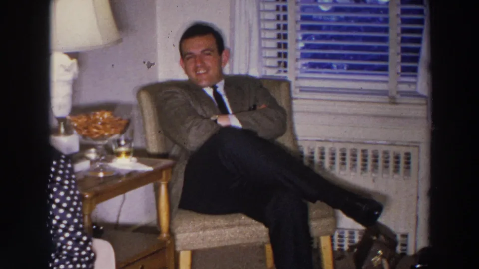 A man in a suit reclines with crossed arms in a Madison living room, seated beside a lamp, snack bowl, and window blinds.