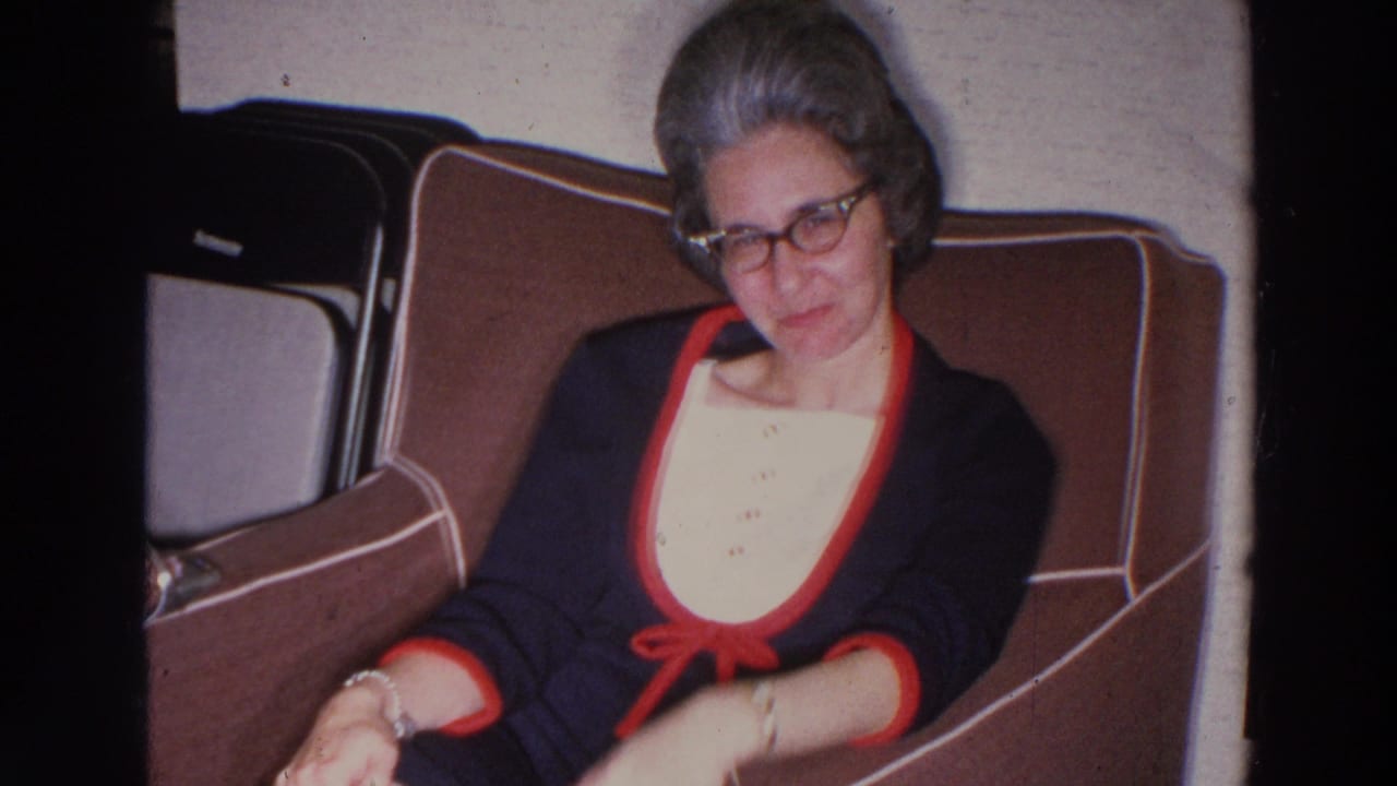 Woman in glasses seated in a brown chair, looking down with one arm moving.