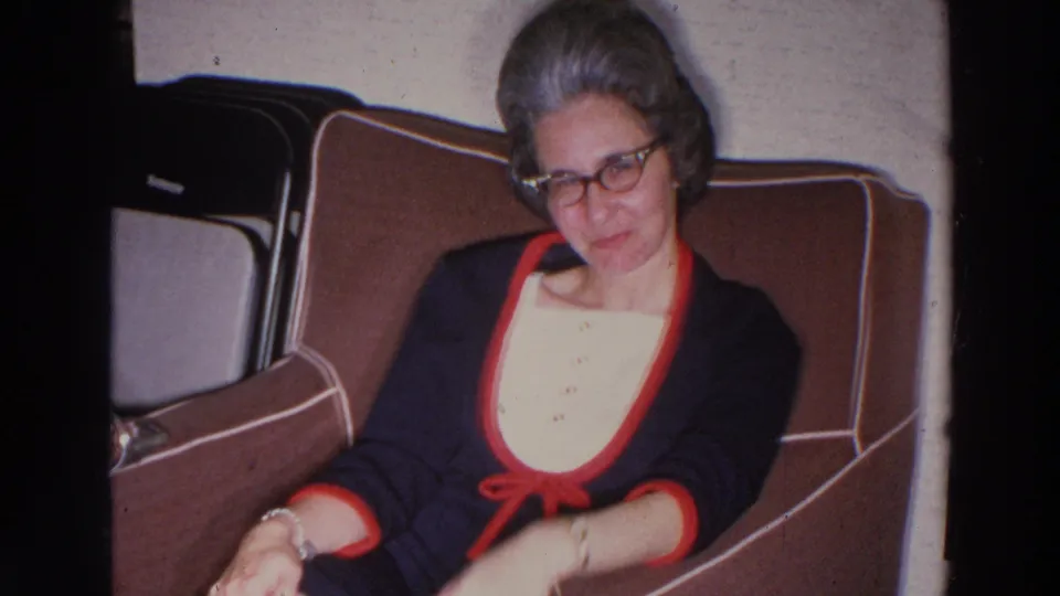 The woman remains seated in the brown upholstered seat, glancing down as her arm moves within the 1966 home-movie frame.
